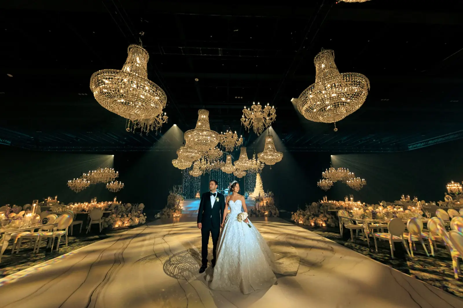 Full wedding production in a Miami ballroom with crystal chandeliers, candlelit reception tables and a bride and groom on a marble aisle
