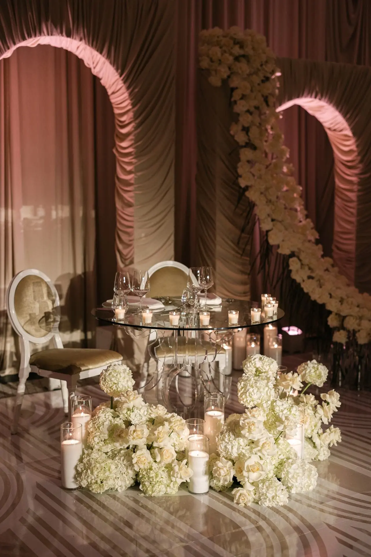 Glass sweetheart table with floor-level white hydrangea and rose cloud against ruched draped arches