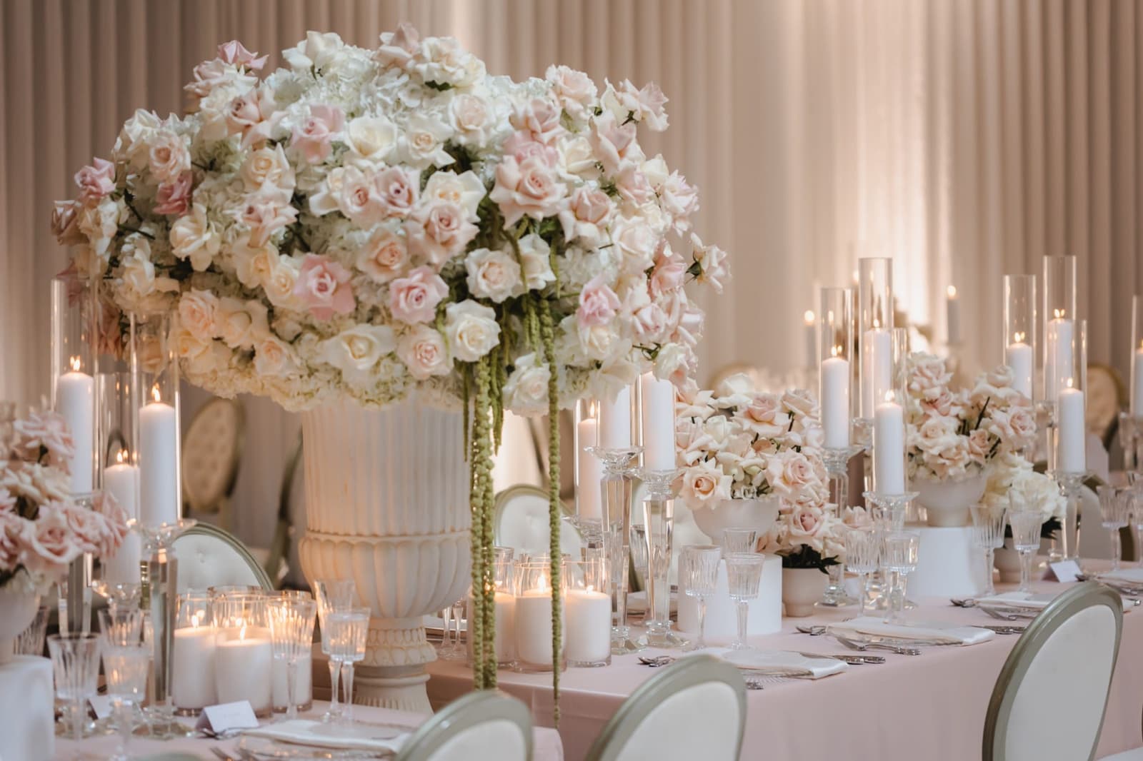 Grand fluted urn overflowing with blush and ivory roses, hydrangea, and amaranthus above candlelit head table at luxury Miami wedding reception
