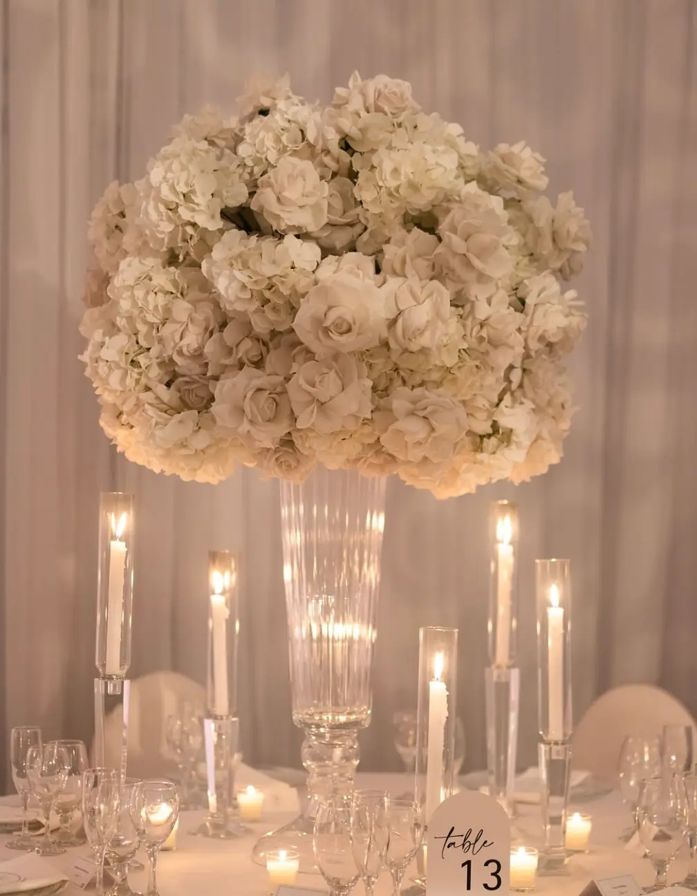 Tall fluted glass trumpet stand with a white hydrangea and rose arrangement above candlelit hurricane tapers at a luxury wedding reception