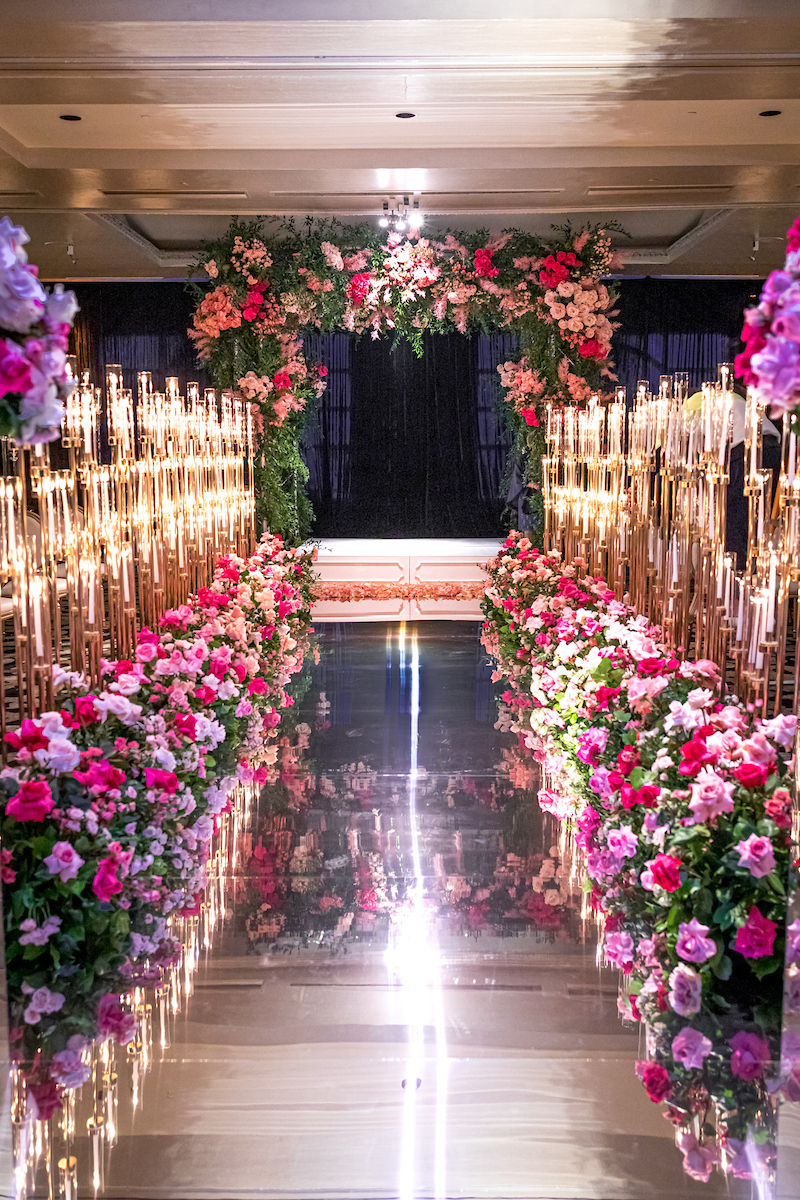 Tall sand-set taper candles in gold holders lining a mirrored aisle with floral arch at a luxury Miami wedding ceremony