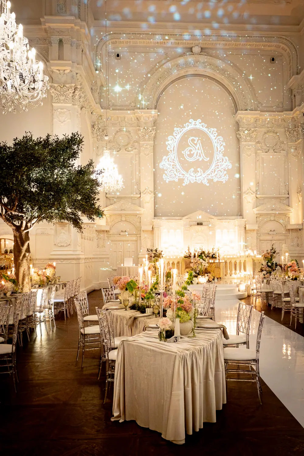 Ornate white ballroom with starry monogram projection, olive trees, and blush candlelit tablescapes at a full production luxury wedding
