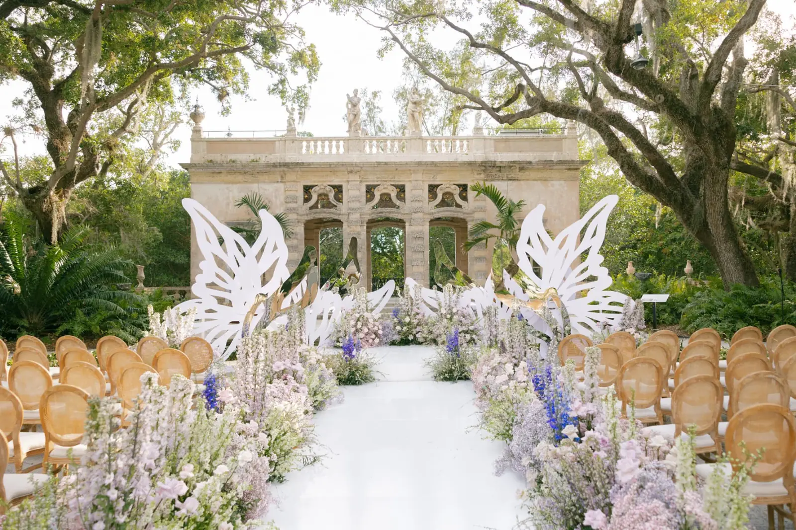 Garden ceremony at Vizcaya with sculptural white butterfly wings, lavender and blush delphinium aisle meadow, and cane chairs at a full production Miami wedding