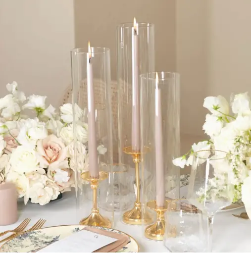 Gold taper candle holders with hurricane glass shades and blush tapers beside white roses on a wedding tablescape