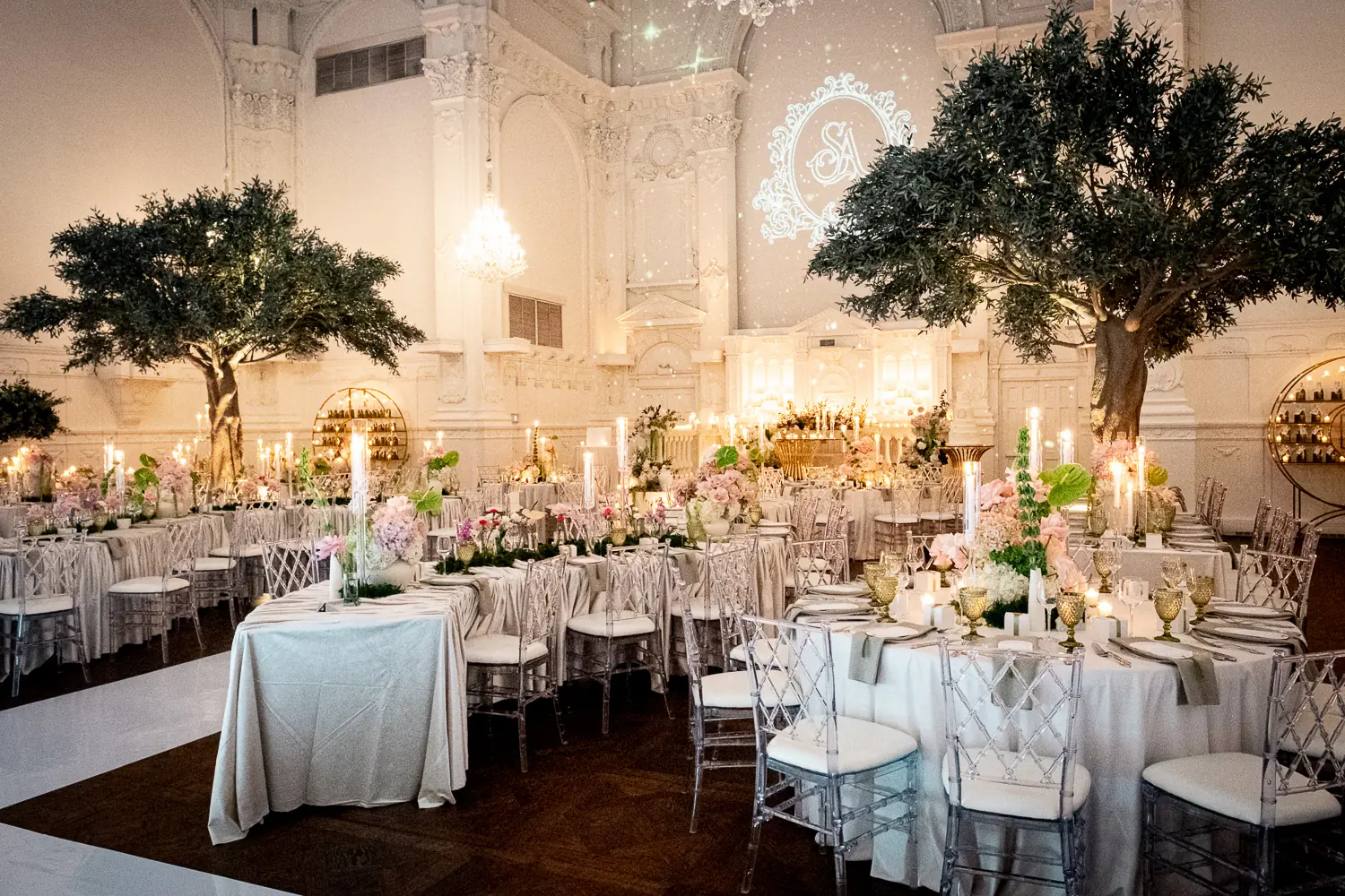 Full-size olive trees, blush and green tablescapes, ghost chairs, and taper candles beneath a starry monogram projection in a white heritage ballroom
