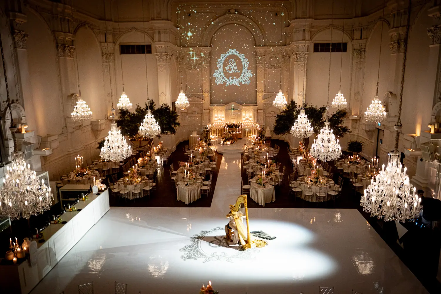 Grand ballroom with rows of crystal chandeliers, a harpist on a white monogram dance floor, and candlelit reception tables at a full event production