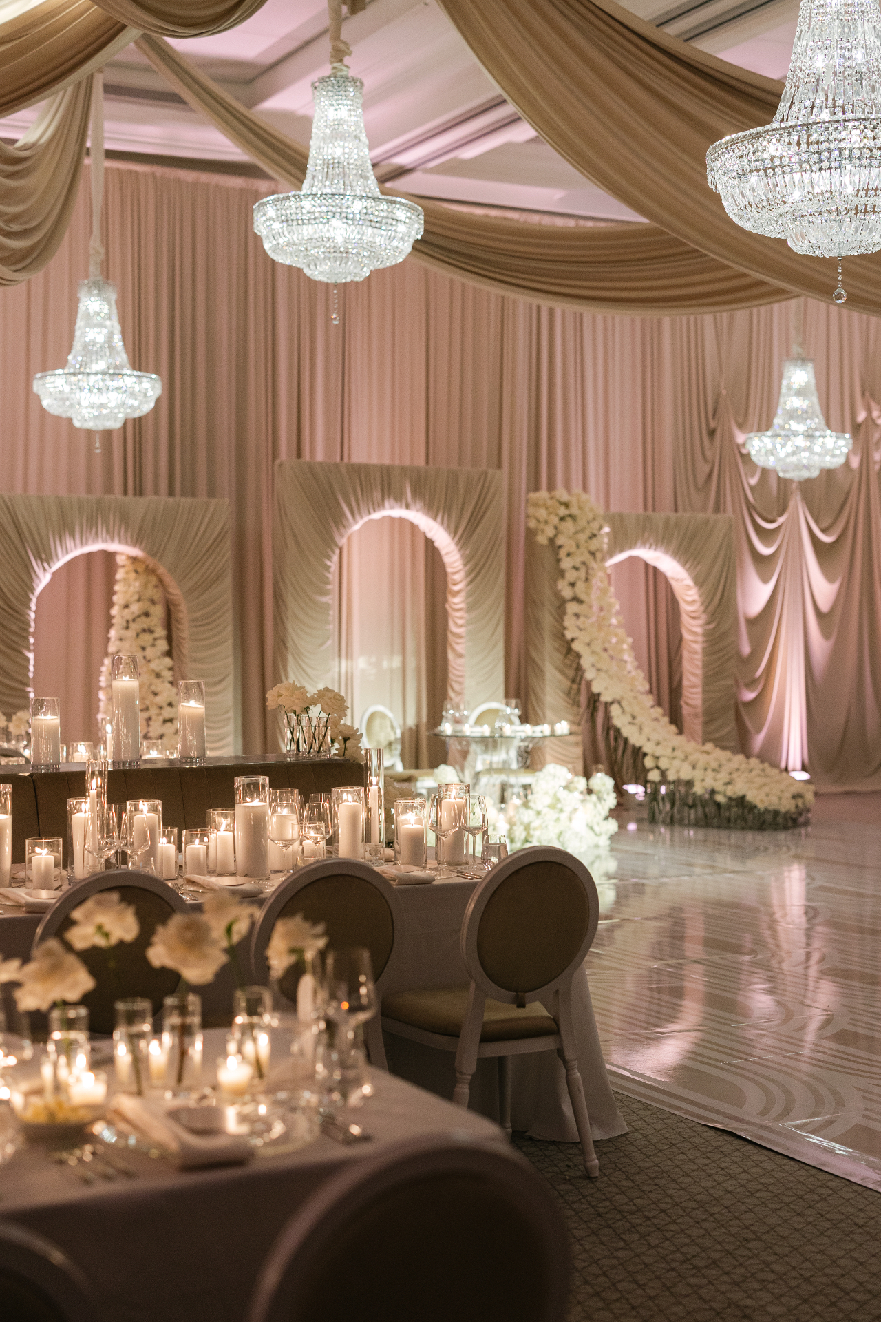 Layered draped arches and crystal chandeliers with cascading white floral installation across a mirrored dance floor