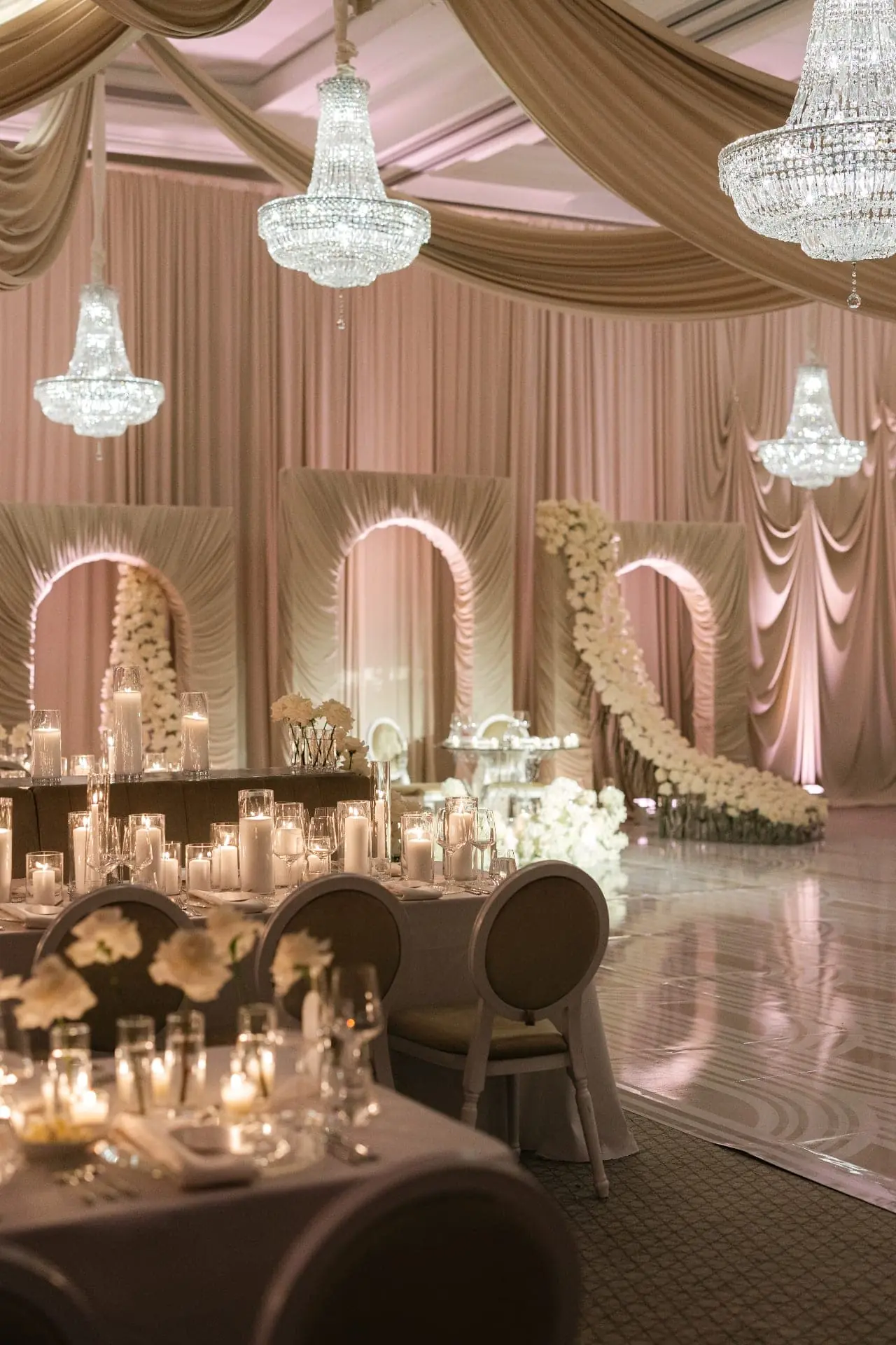 Layered draped arches and crystal chandeliers with cascading white floral installation across a mirrored dance floor