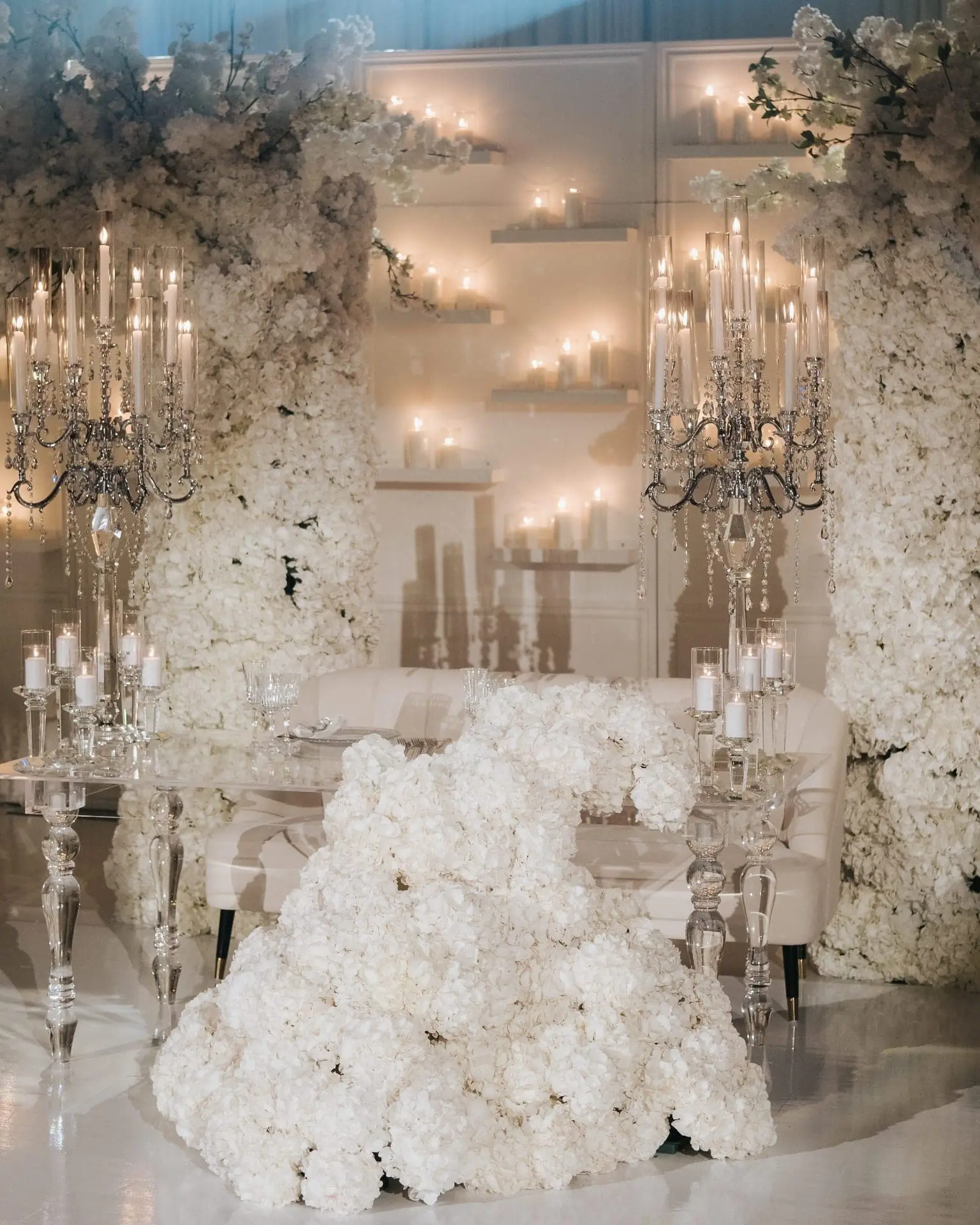 All-White Hydrangea Sweetheart - All-white hydrangea sweetheart table with towering crystal candelabras, cascading floral pillars, and candlelit votive wall by Aura Design Flowers Miami