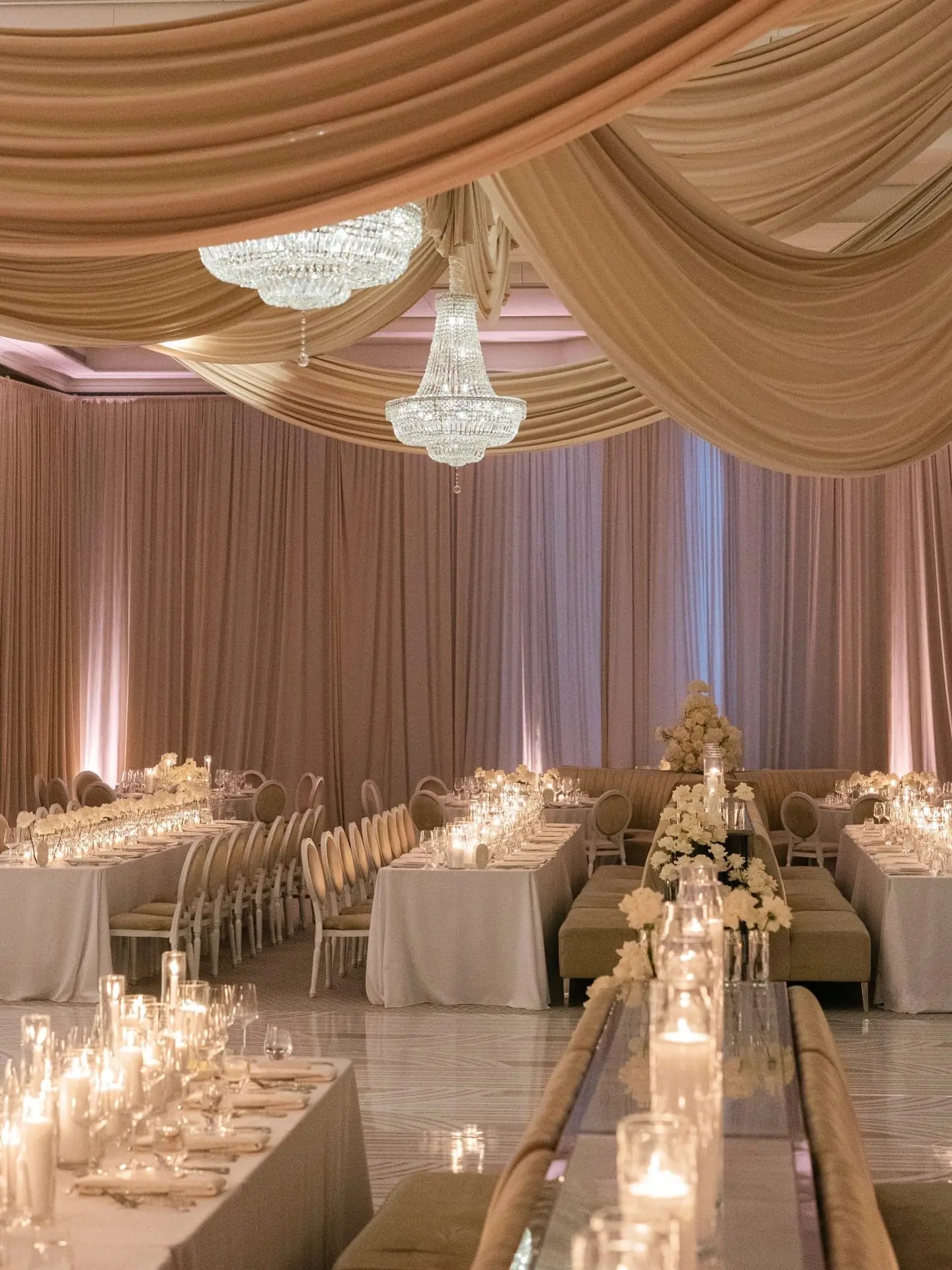 Blush and champagne draped ballroom with crystal chandeliers above mirrored long tables and candlelit white florals at luxury Miami wedding