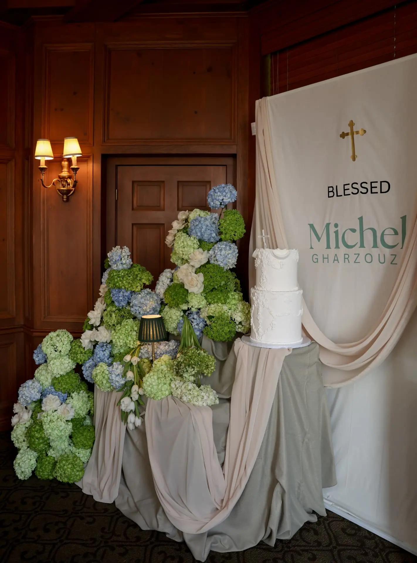 Baptism cake table with cascading blue and green hydrangea, white roses, and blush draped backdrop with gold cross signage