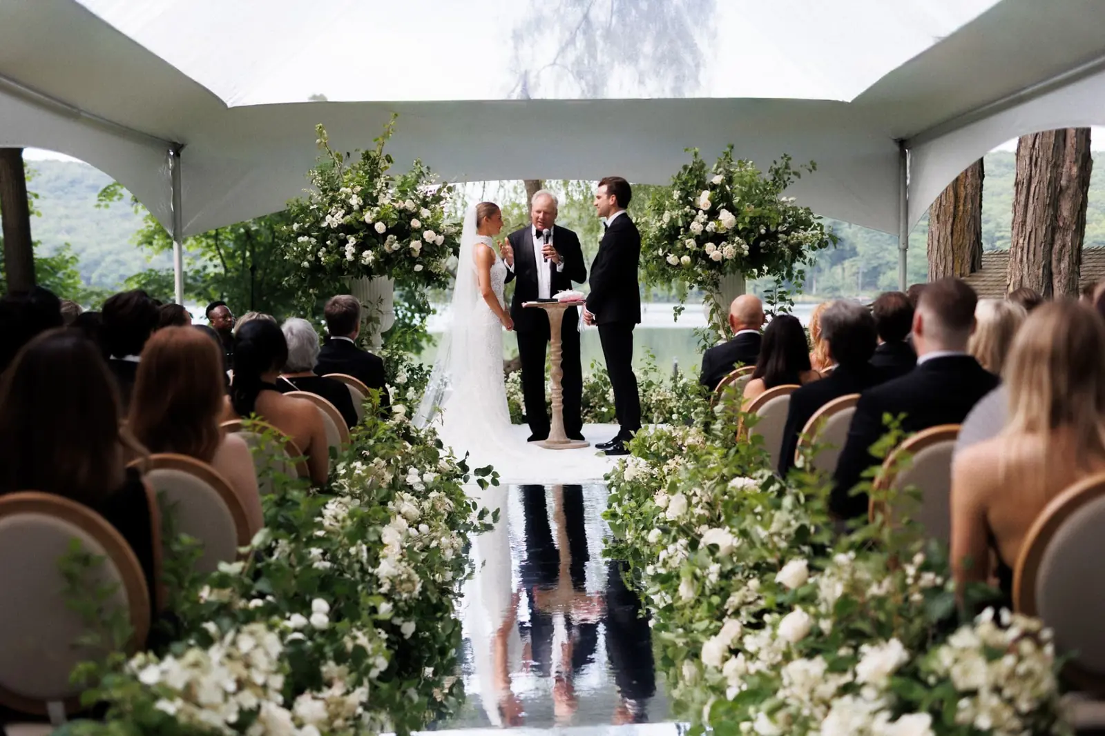 Tented Lakeside Ceremony - Tented lakeside wedding ceremony with towering white rose and greenery urns, lush aisle meadows, and a mirrored aisle runner by Aura Design Flowers Miami