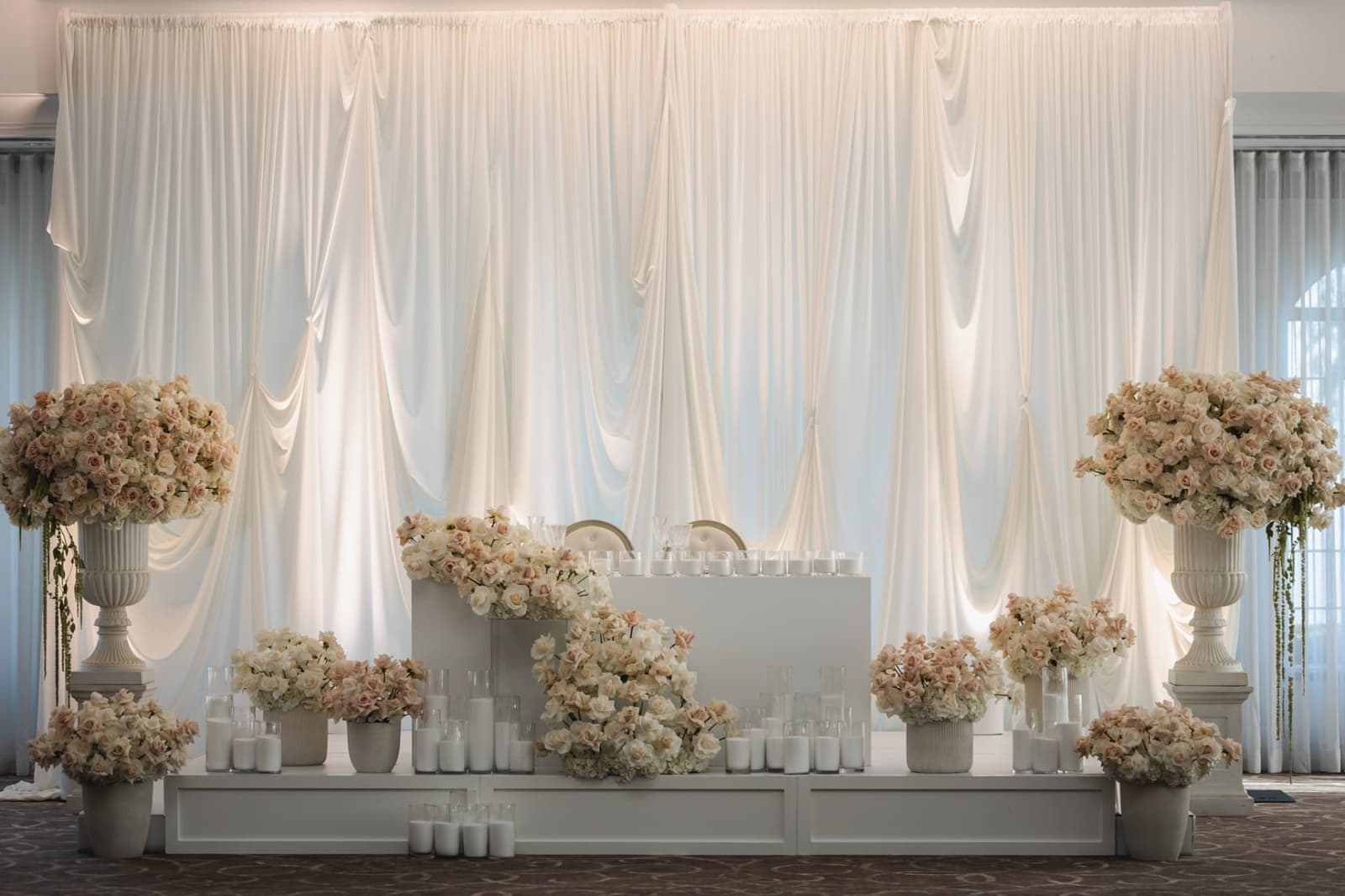 Towering blush and ivory rose urns flanking white draped backdrop with cascading rose sweetheart stage and pillar candles at luxury Miami wedding