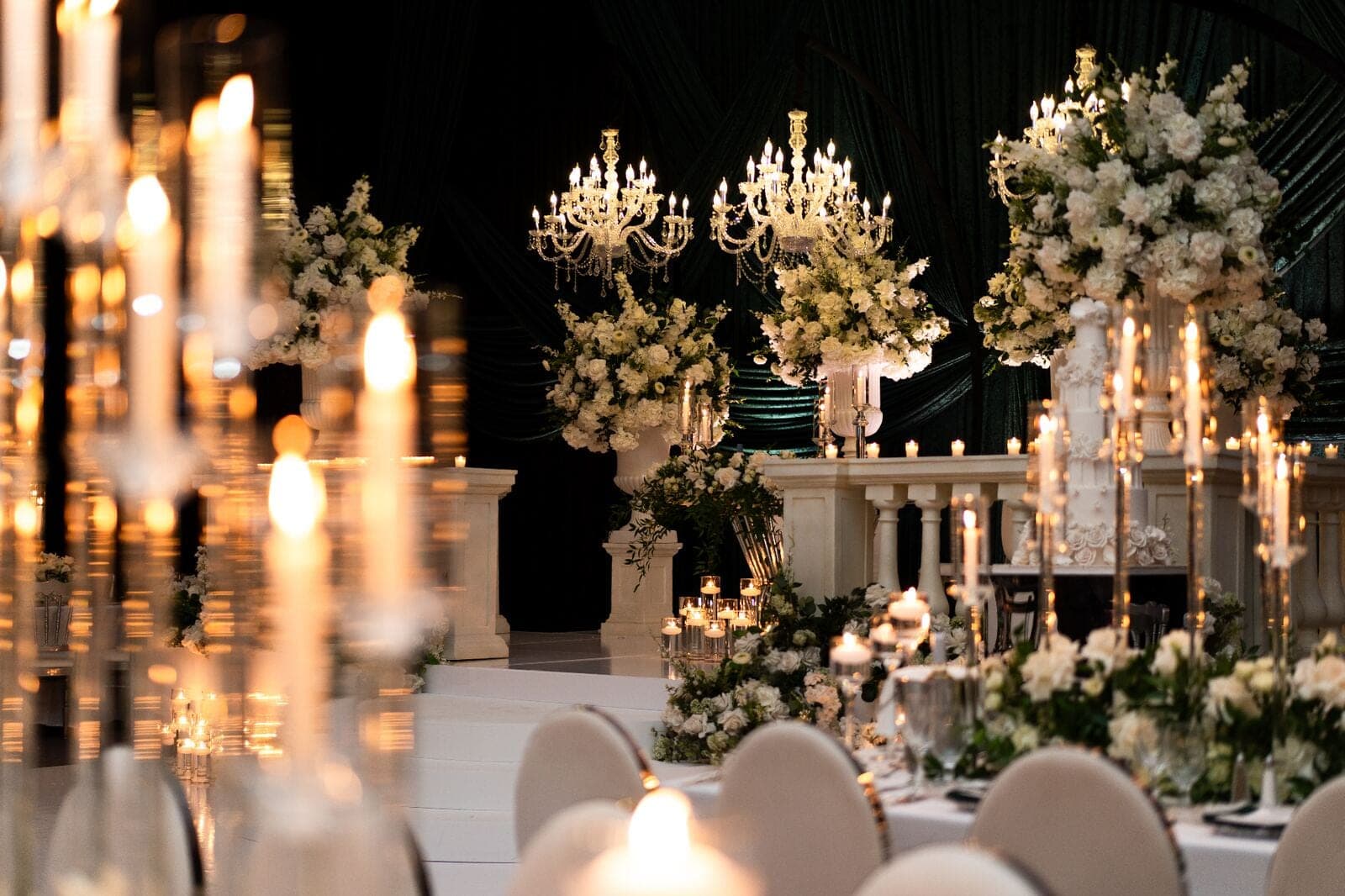 Candlelit ballroom wedding with towering white floral pedestals, crystal chandeliers, and emerald draped backdrop in Miami