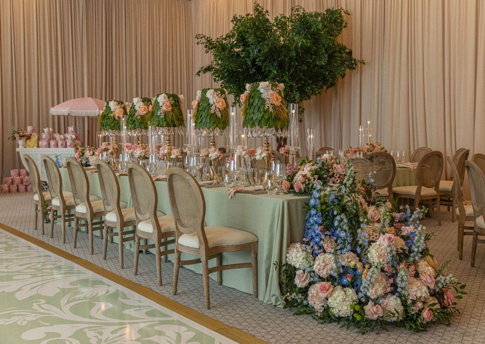 Sage green head table with row of leaf-and-rose lampshade centerpieces, pastel floral cascade, and topiary tree at luxury Miami baby shower