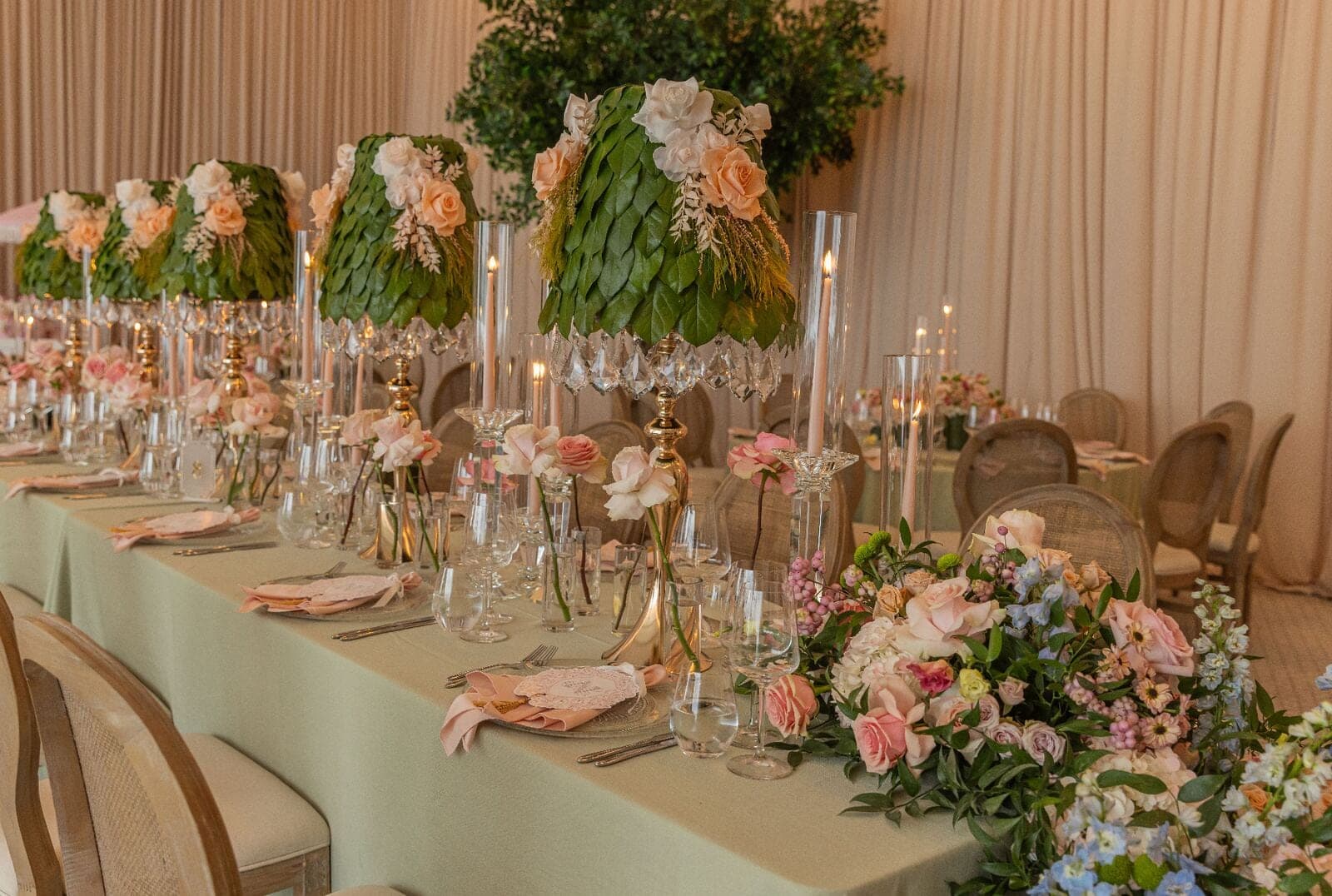 Row of green leaf lampshade centerpieces with peach roses and crystal candleholders along sage tablescape at Miami baby shower