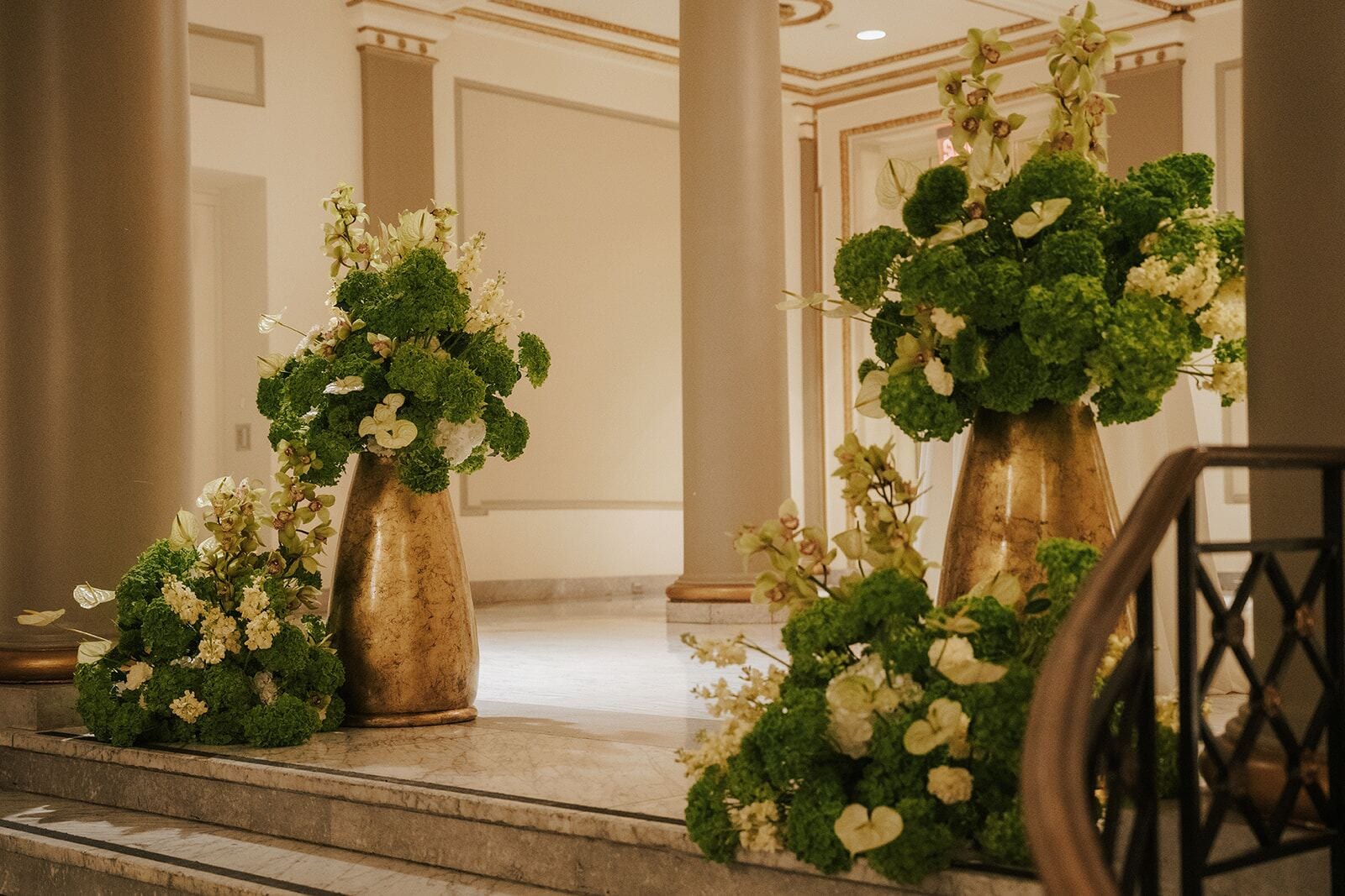 Grand Staircase Vases - Gold vessels overflowing with green and ivory blooms on a marble staircase by Aura Design Flowers Miami